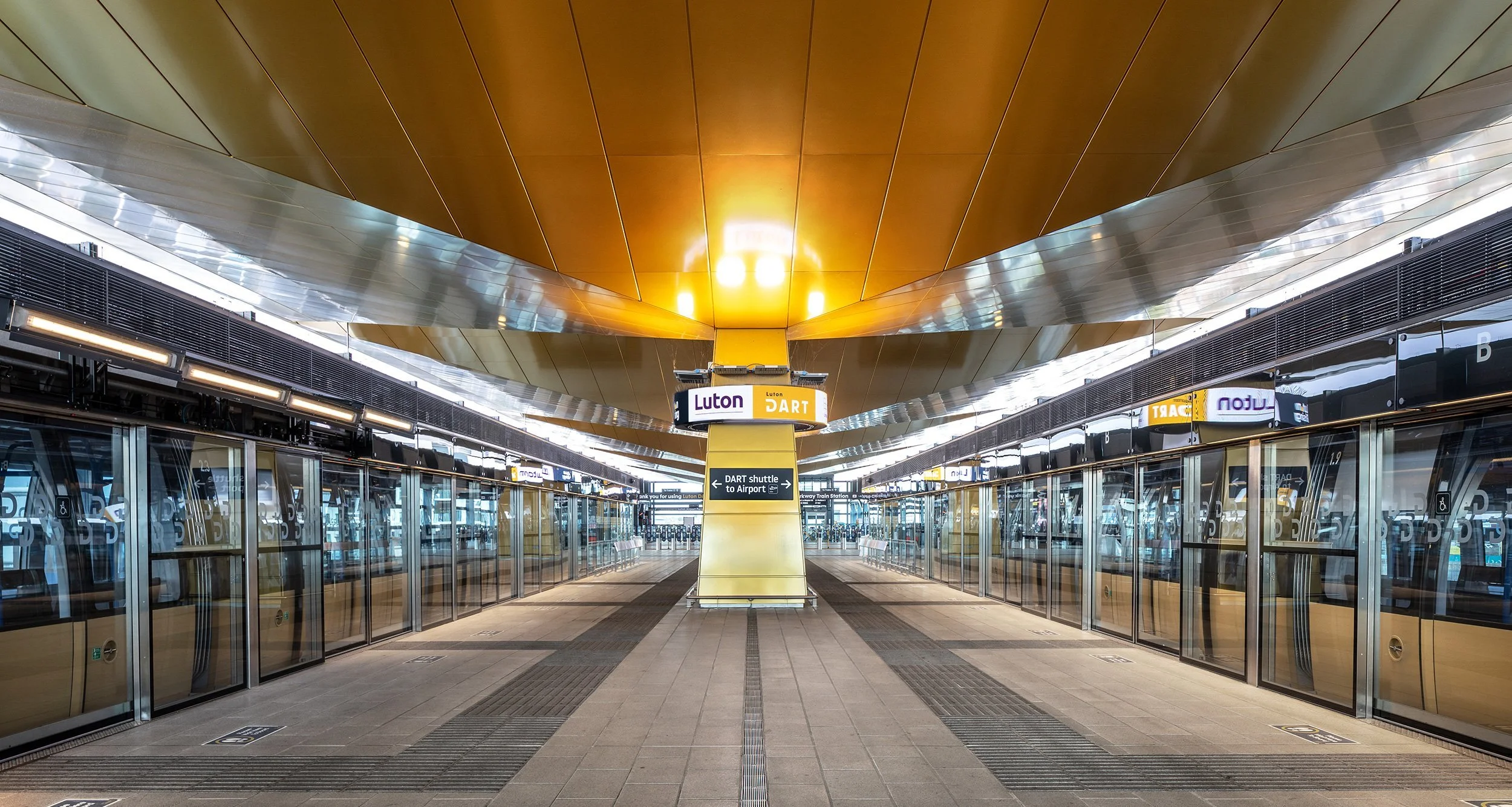 Overhead exit signs in a pale stone ticket hall, arrows pointing toward rail, taxi, and city centre, a row of unused ticket machines along the left wall, and a traveller looking up from a paper boarding stub, 1600x1000