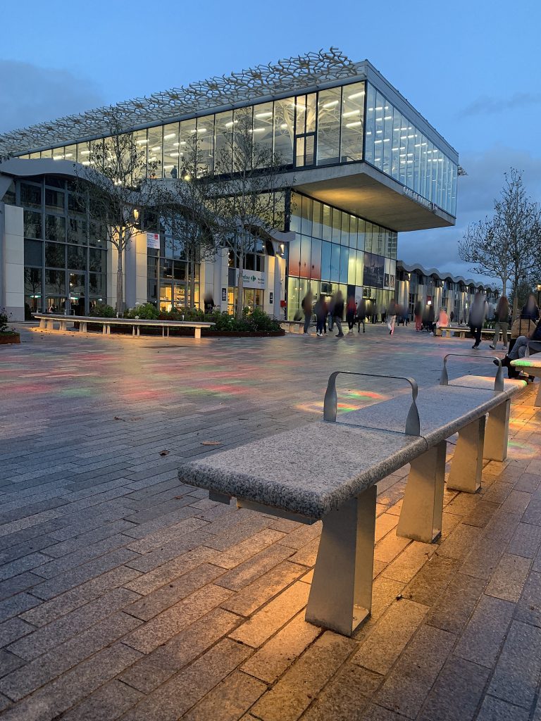Station forecourt at dusk with wet paving reflecting amber lamps, a covered taxi lane to the right, a bus shelter with a printed timetable, and one traveller pausing under a clock with a backpack and a folded paper map, 1600x1067